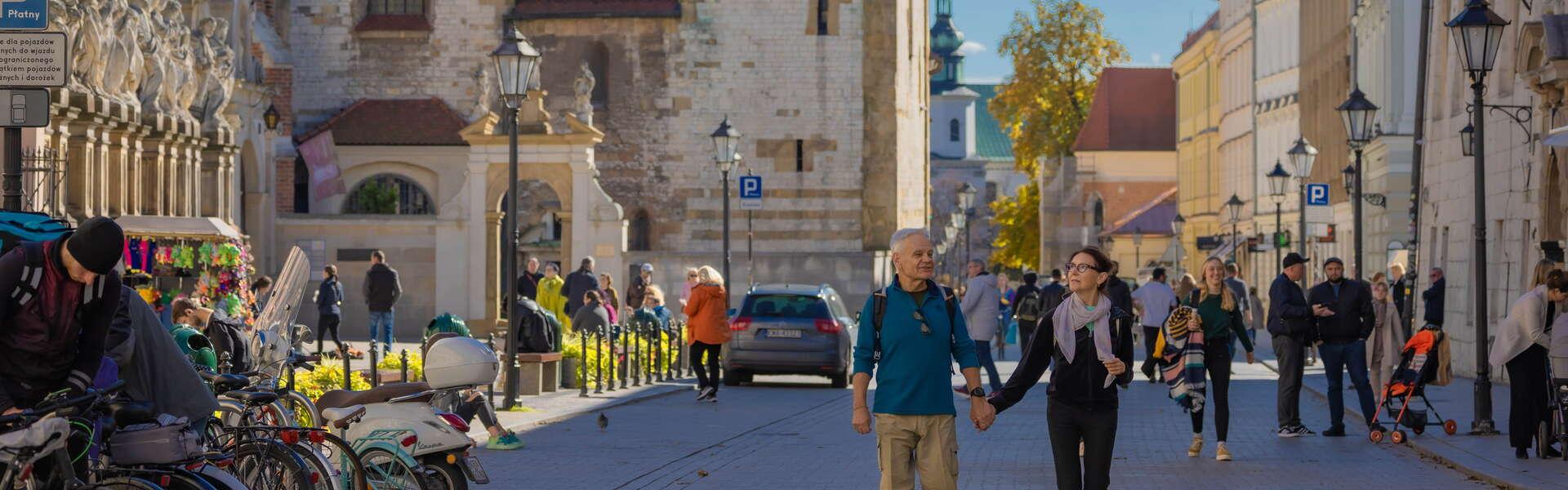 Ein Mann und eine Frau halten sich an den Händen und gehen in Kraków spazieren. Links und rechts Stadthäuser. Im Hintergrund ein Kirchturm.