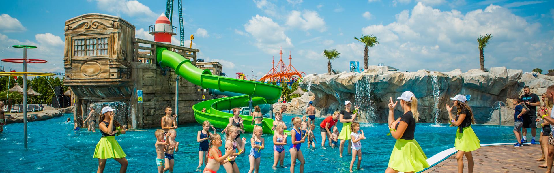 Two female dance instructors  on the edge of the paddling pool keep an eye on young children dancing. Behind them is a structure with a lighthouse on top and a winding slide. In the background is a tall structure and next to it on the right are rocks with artificial palm trees and a waterfall. On the left, there is a fountain and a serene sky.